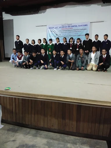 Students in uniform seated and standing on the auditorium stage beneath a competitive examination banner.